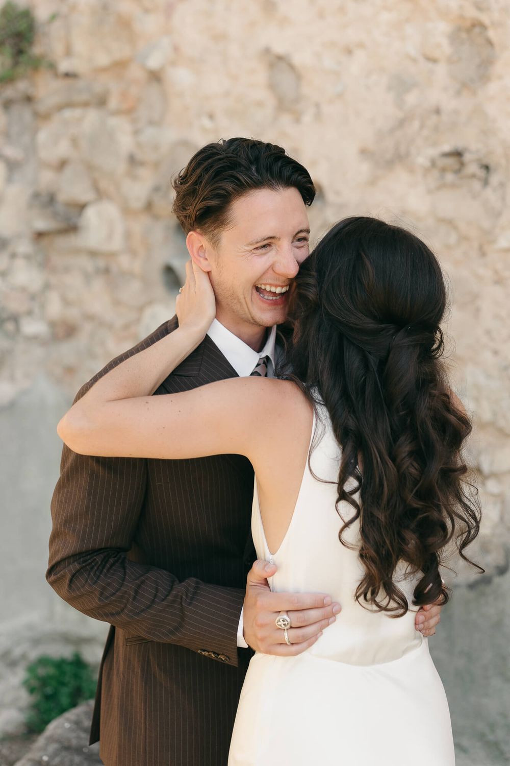 Joyful groom in brown suit laughing as bride in white dress embraces him outdoors near stone wall.