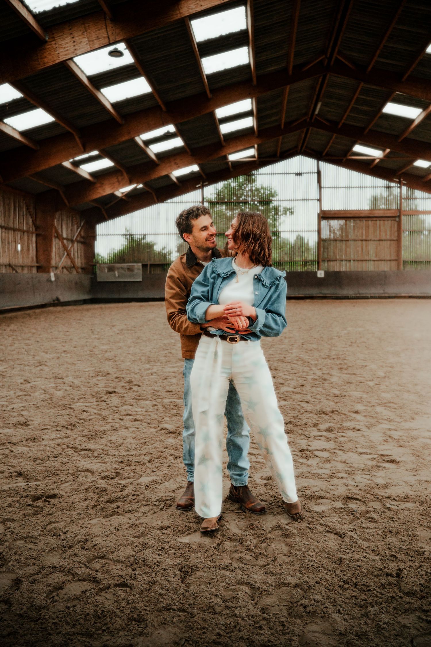 Couple embracing and smiling in a sandy indoor riding arena, wearing casual denim and brown jacket outfits.
