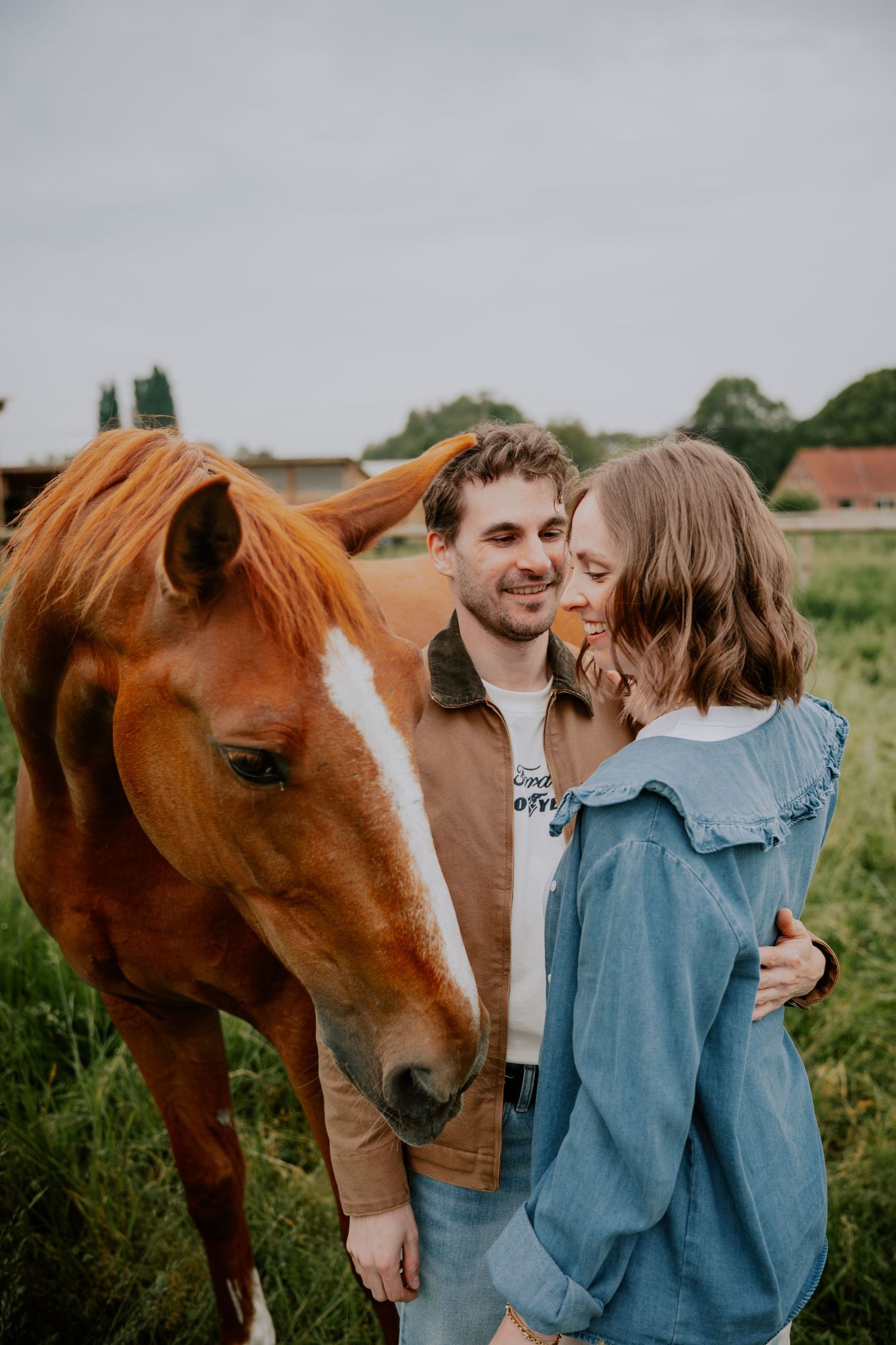 A couple shares a tender moment beside a chestnut horse in a green field, the man in brown jacket embracing the woman in denim.