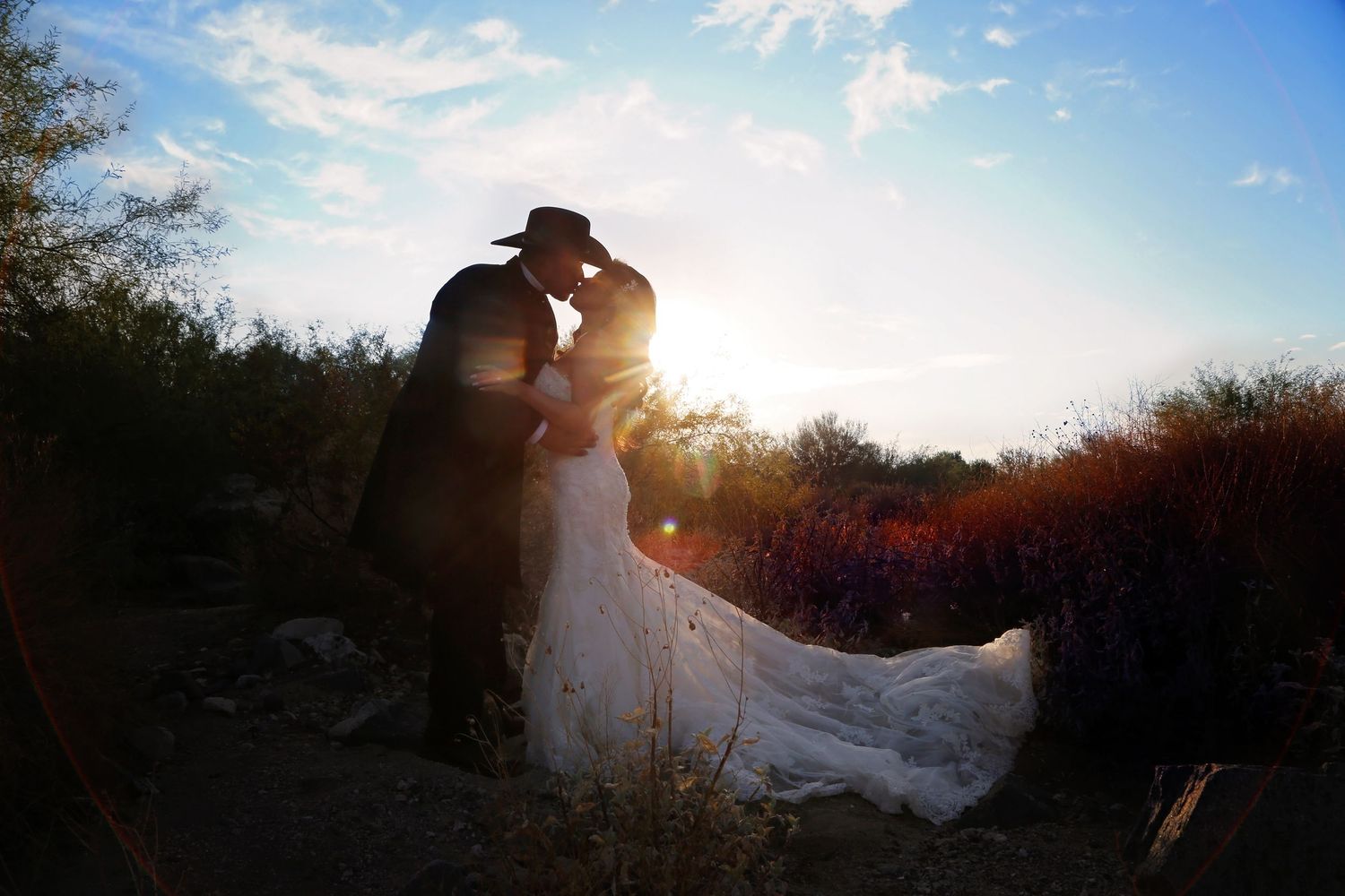 A groom in a cowboy hat kisses his bride in a flowing white wedding gown as the sun sets behind desert brush.