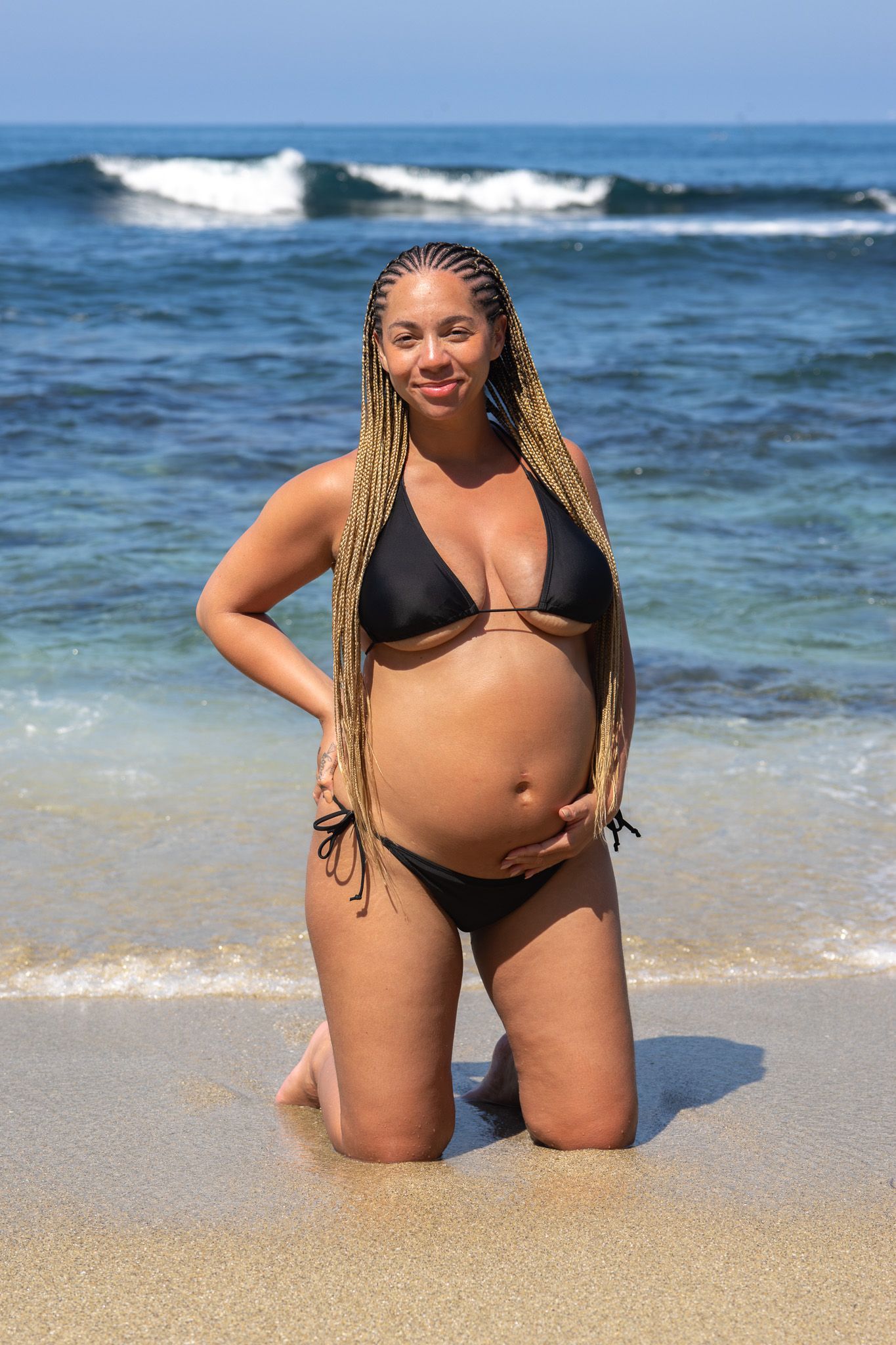 San Diego beach maternity photography featuring a pregnant woman in a black bikini kneeling on the sand by the ocean waves.