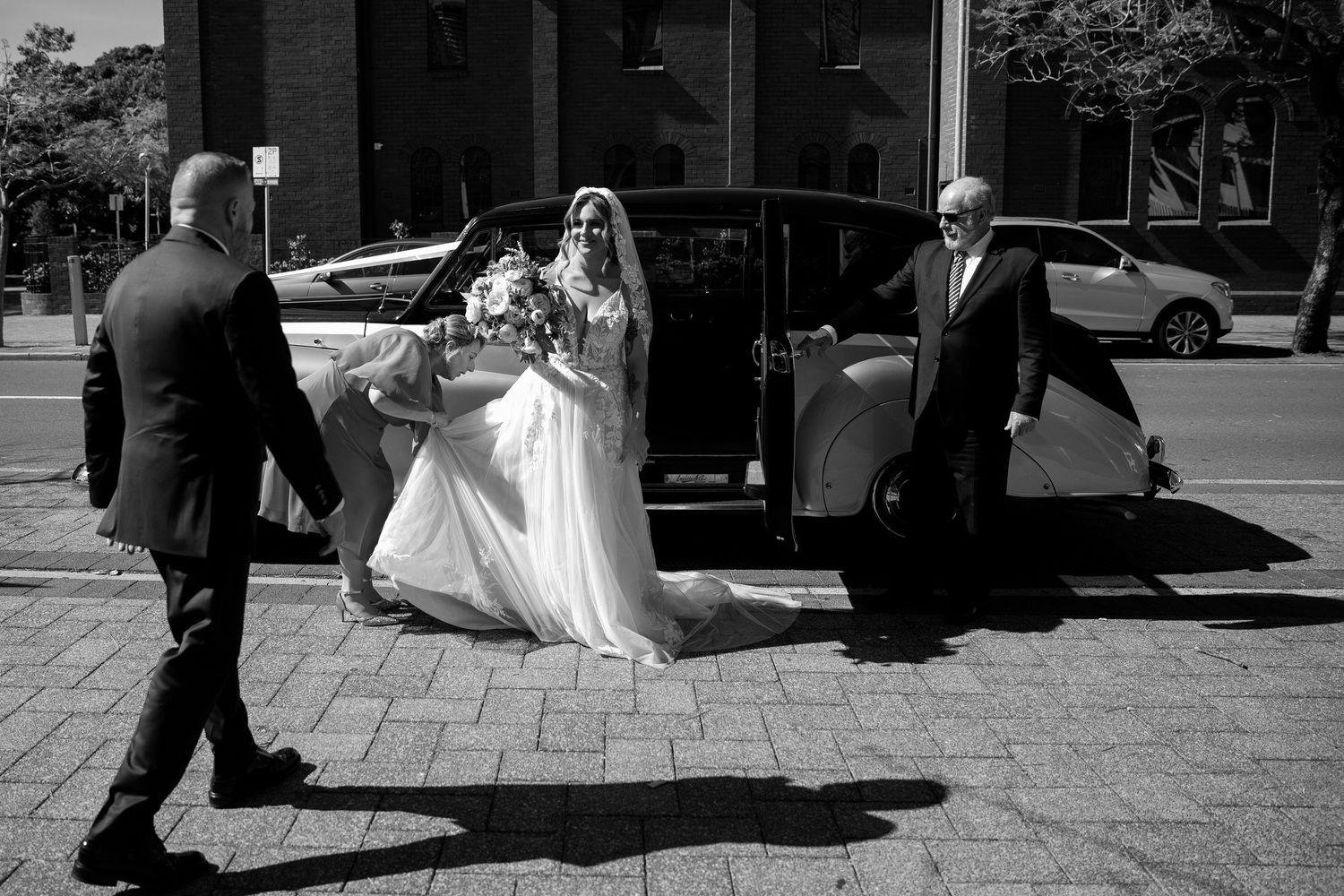 Black and white photo of a bride in a white gown exiting a black car, assisted by two men on a brick sidewalk.