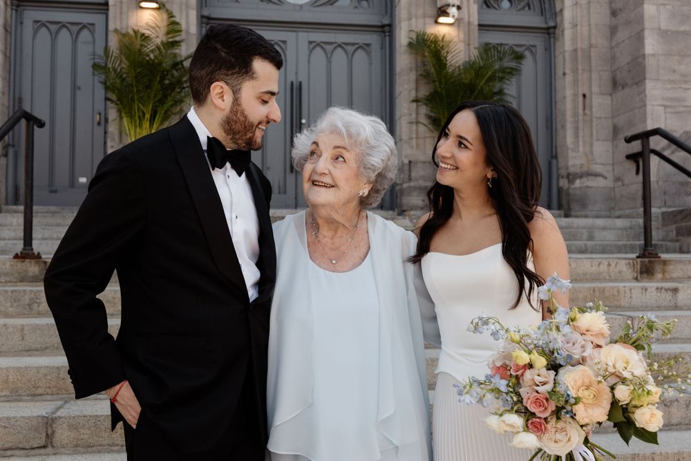 Bride and groom pose with elderly grandmother on church steps during wedding celebration.