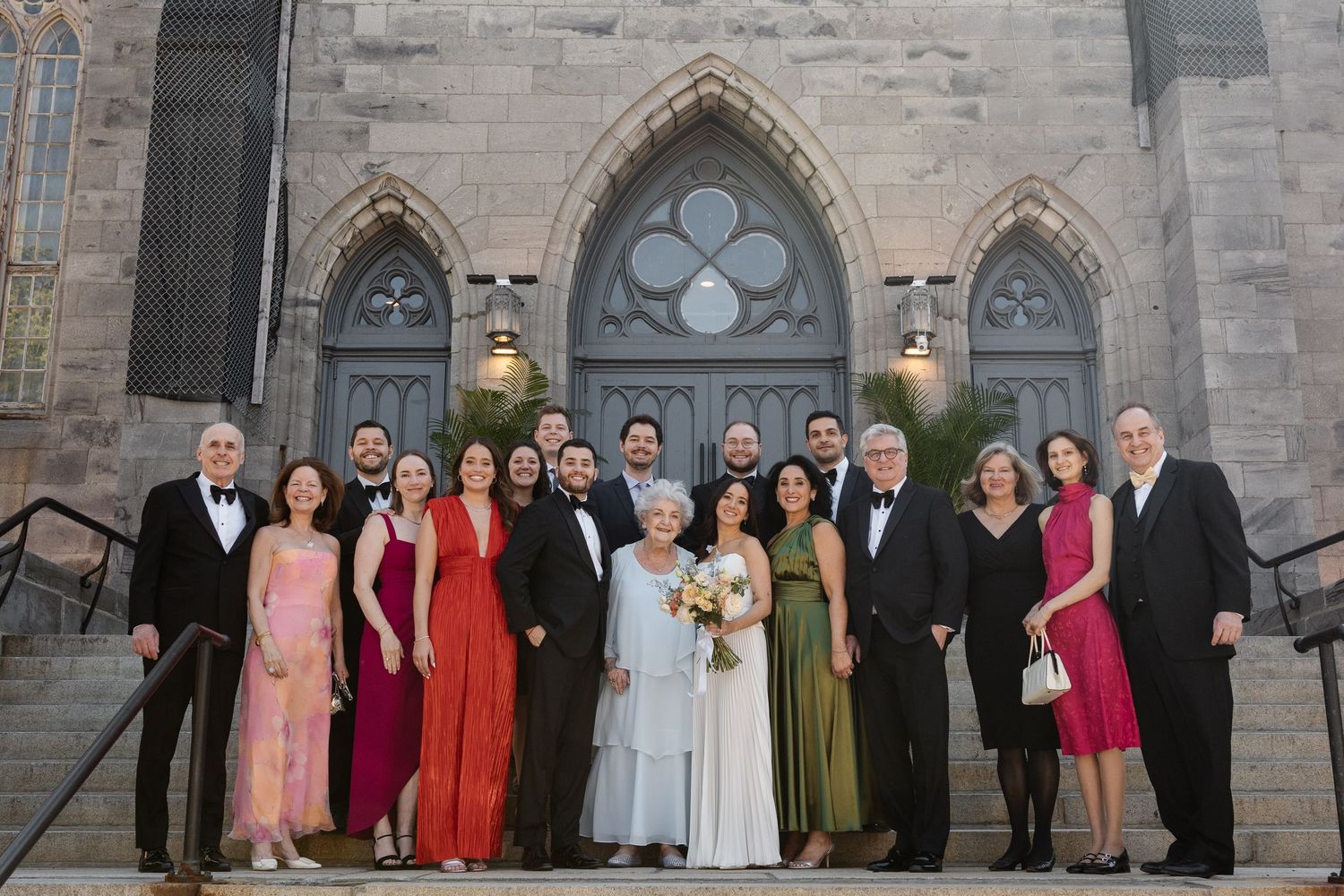 Wedding party posing on church steps in formal attire, featuring bride in white gown with gothic stone architecture backdrop.
