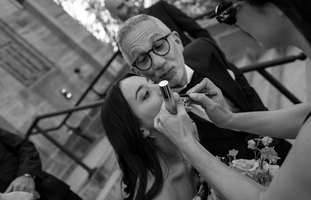 Black and white photo of a bride getting lipstick applied by a makeup artist outdoors at a wedding.