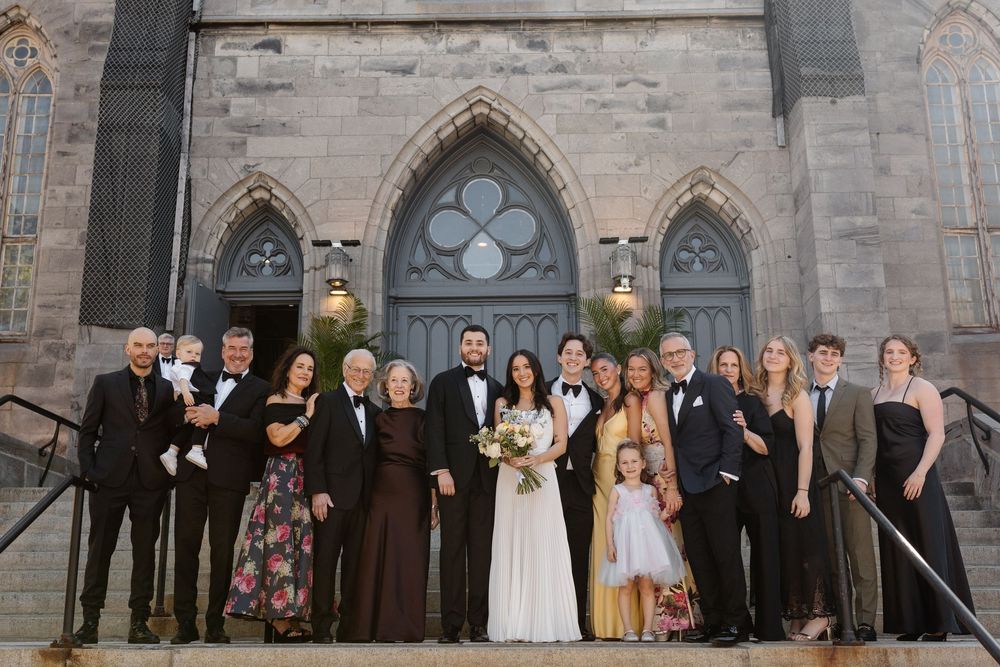 Wedding party posing on church steps in formal attire, bride in white gown holding bouquet, stone Gothic architecture backdrop.