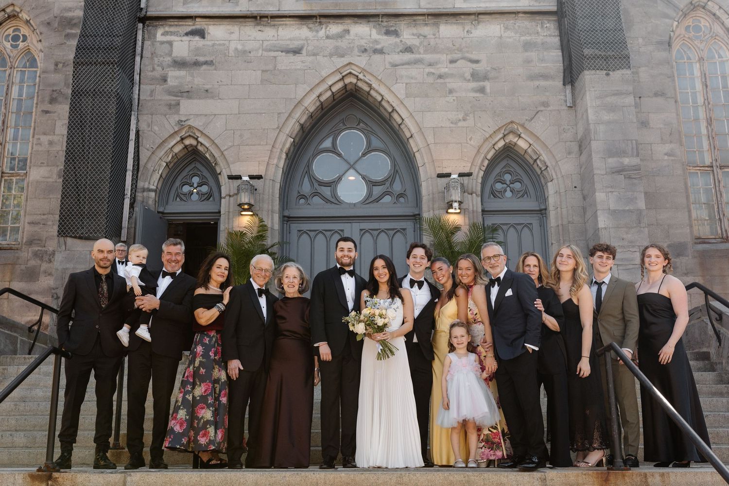 Wedding party posing on church steps in formal attire, bride in white gown holding bouquet, stone Gothic architecture backdrop.