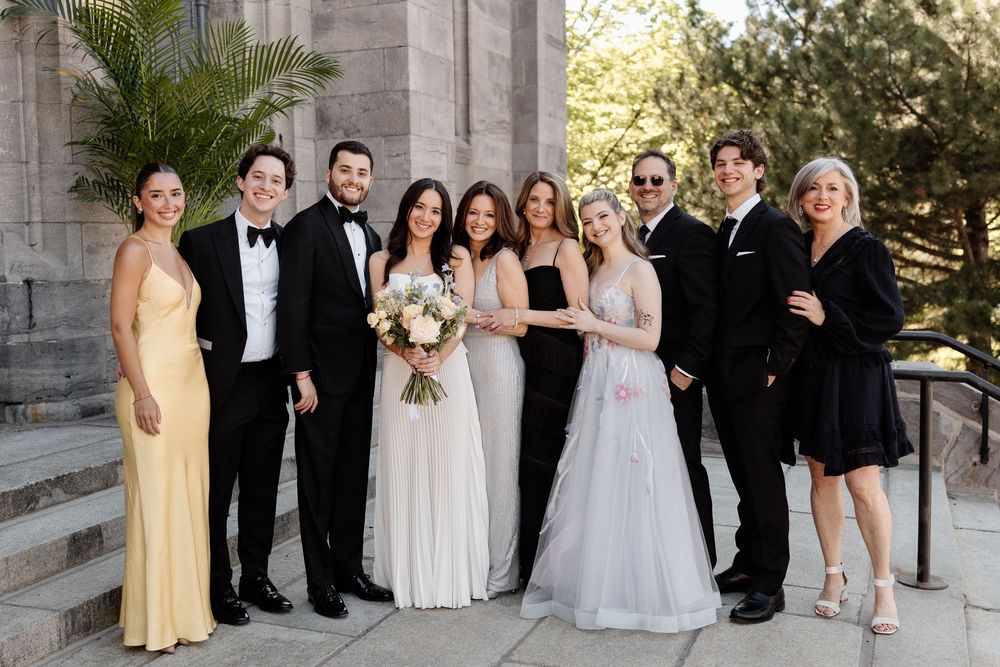 Wedding party group photo with bride, groom, and guests in formal attire outdoors.