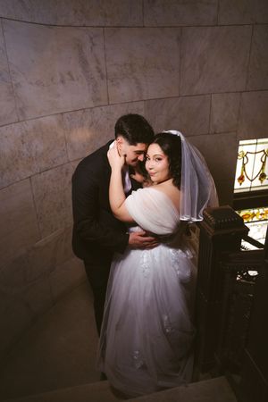 Bride in white gown and veil embraces groom in black suit against marble wall in romantic wedding portrait.