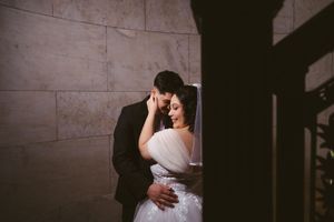 A bride and groom share a romantic embrace against a marble wall in soft dramatic lighting on their wedding day.