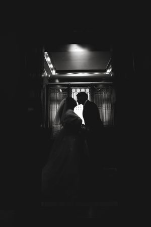 Black and white silhouette of a bride and groom sharing a romantic moment in a dimly lit elegant venue.