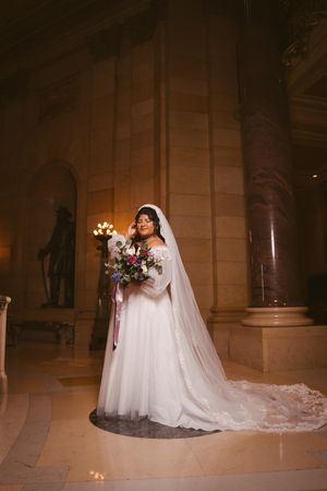 Bride in off-shoulder white gown with long veil holding floral bouquet in ornate marble building interior.