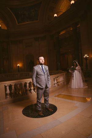 Groom in gray suit standing on marble floor of grand historic building with bride visible in background.