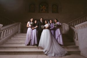Wedding party poses on grand staircase, bride in white gown with bridesmaids in lavender dresses and groom in black suit.