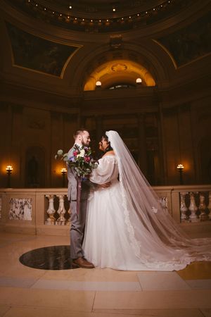Bride and groom share romantic moment in ornate rotunda, her flowing veil trailing on marble floor.