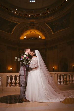 Bride and groom share a kiss inside an elegant domed hall with ornate architecture and warm golden lighting.