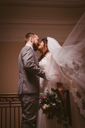 Bride and groom share a romantic moment on staircase, veil flowing, as bride holds purple floral bouquet.