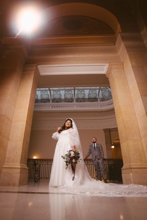 Bride in white gown and veil holds bouquet inside elegant wedding venue with arched ceilings as groom stands behind.
