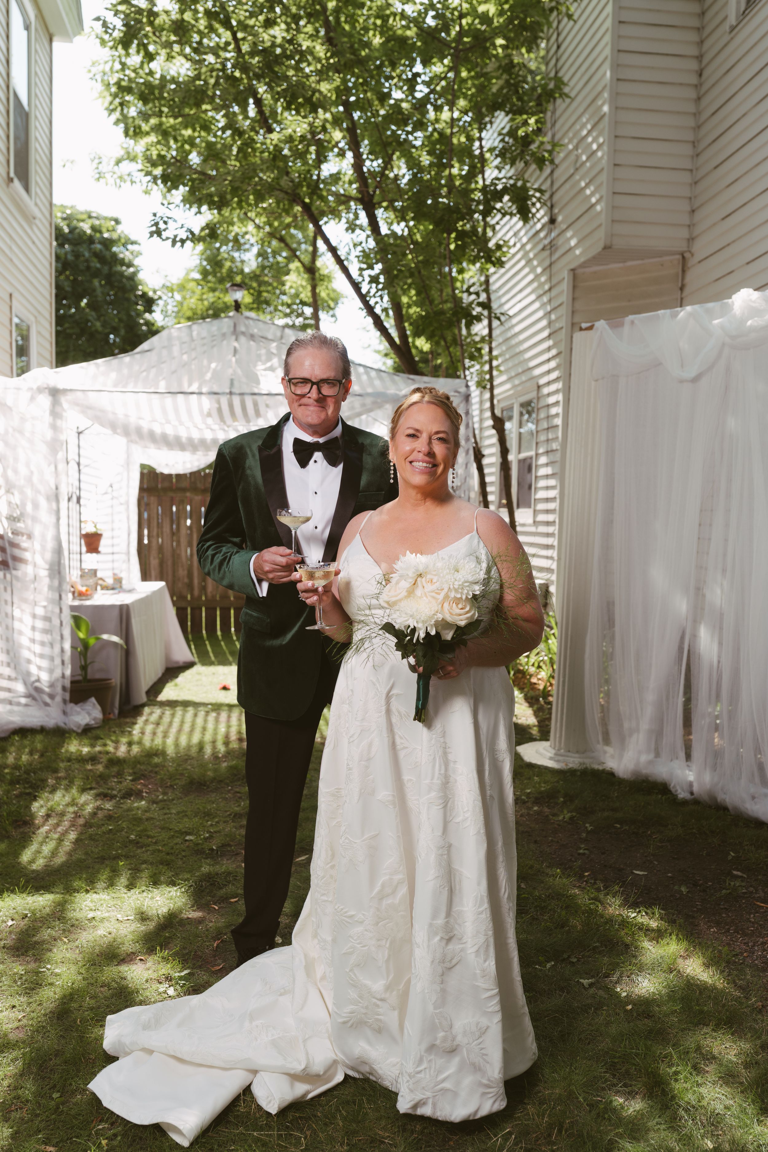 Bride in white gown holding bouquet stands with groom in green velvet tuxedo toasting champagne in backyard wedding.