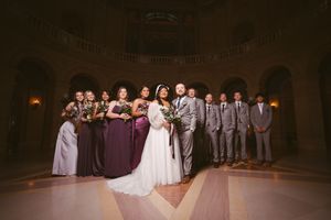Wedding party poses in elegant venue, bridesmaids in purple gowns and groomsmen in gray suits flanking bride and groom.