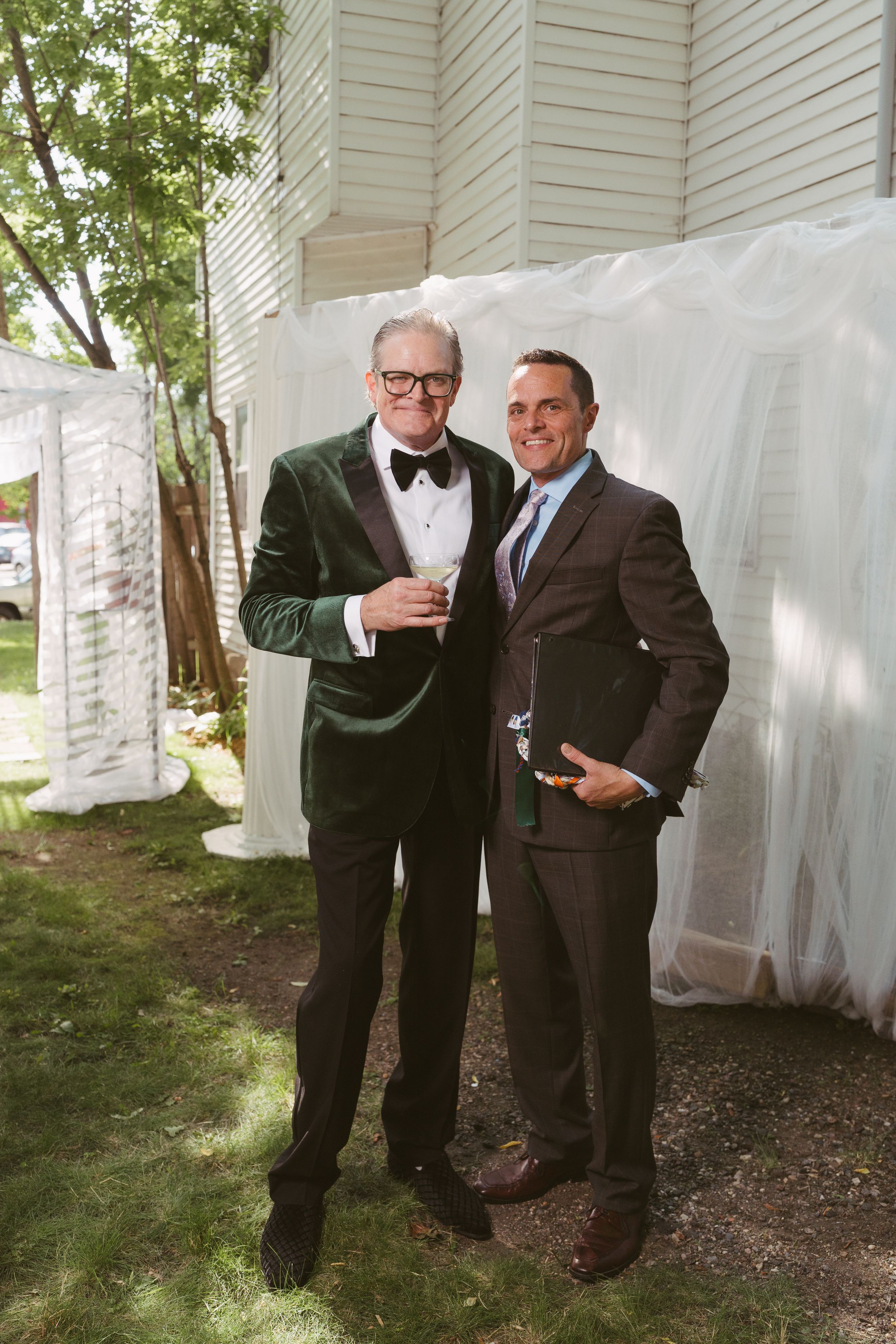 Two men in formal suits smile together at outdoor wedding reception, one wearing green velvet tuxedo with bow tie.