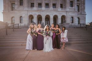 Bride with bridesmaids in purple and gray gowns posing on steps of a grand stone building at golden hour.
