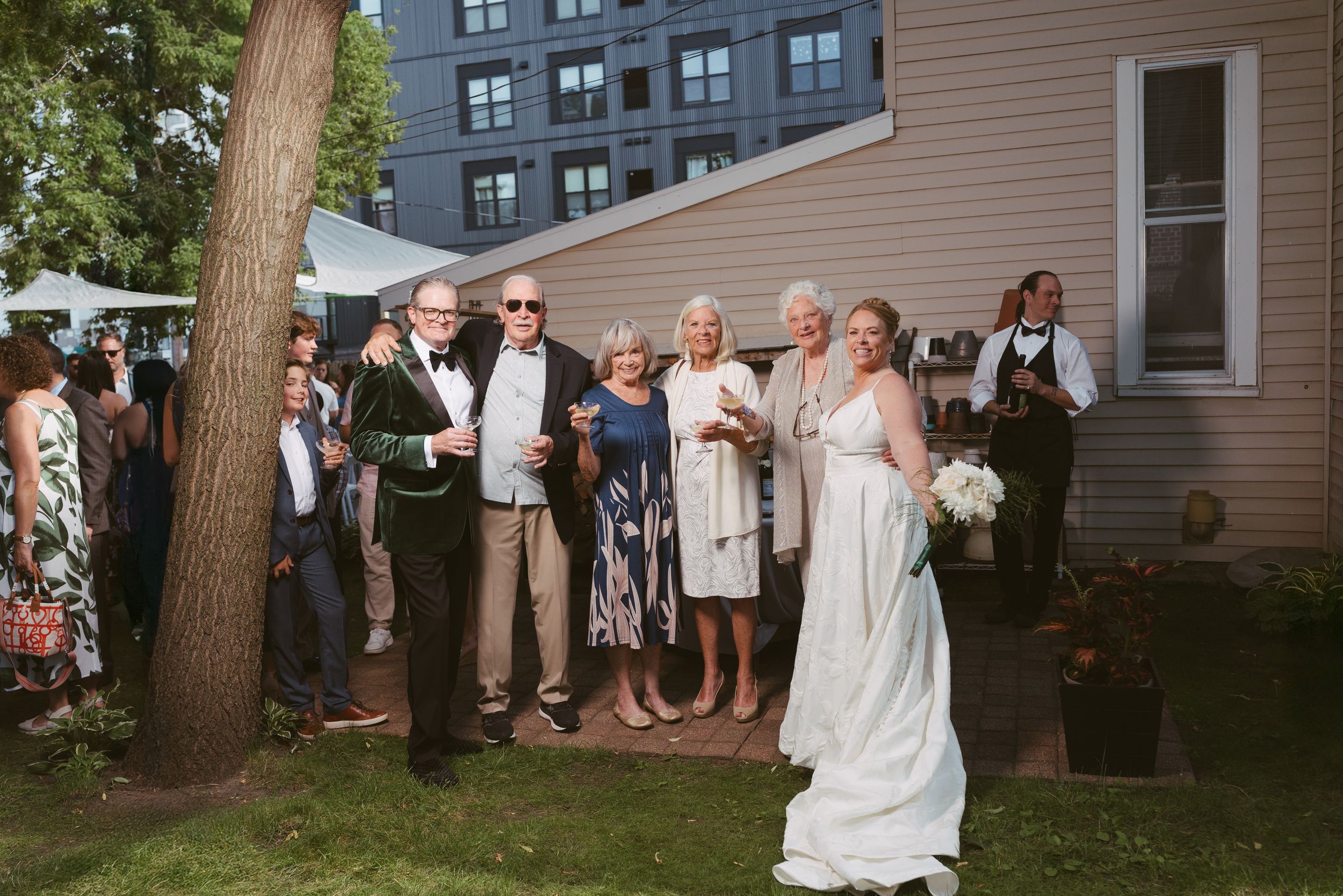 Bride in white gown holds bouquet while guests gather outdoors during a backyard wedding ceremony.