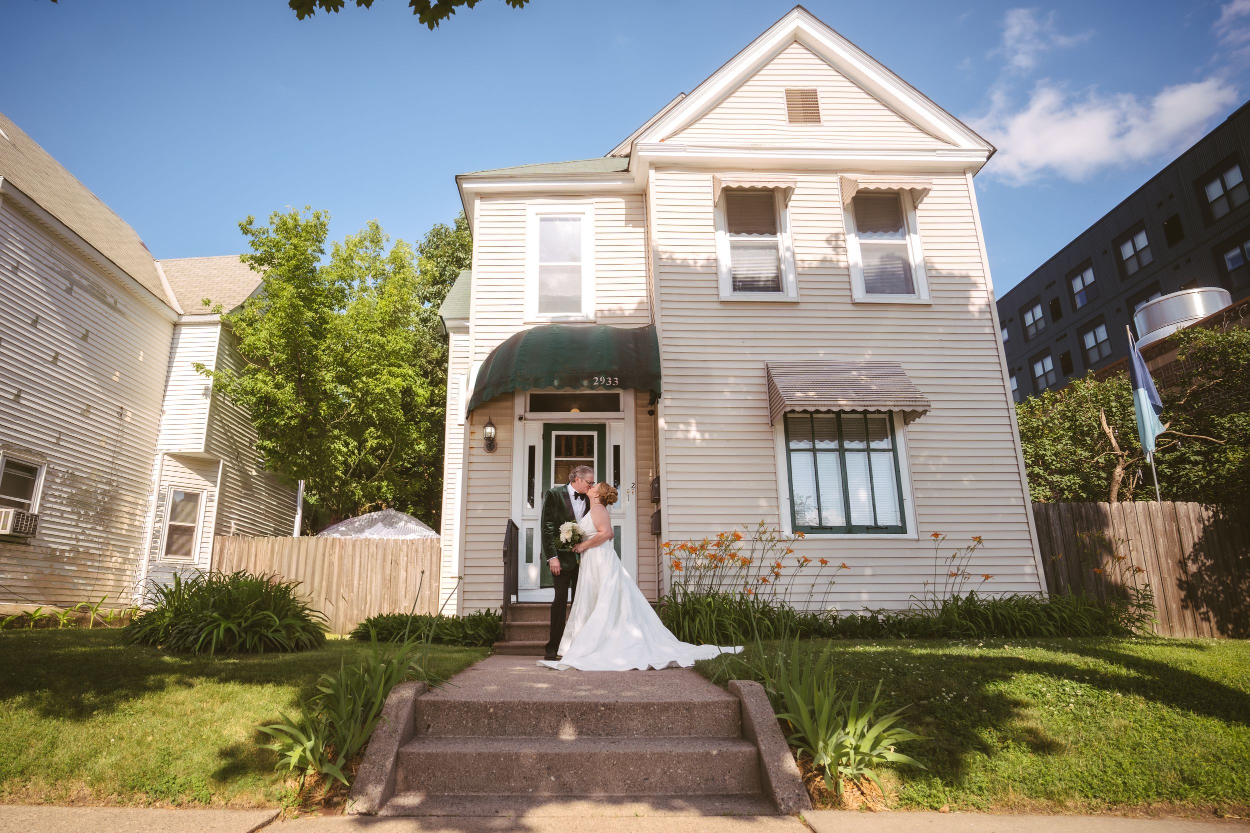 Bride and groom pose in front of a charming white two-story house on their wedding day.