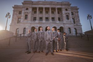 Groom and groomsmen in gray suits pose on steps of a grand stone building at golden hour.