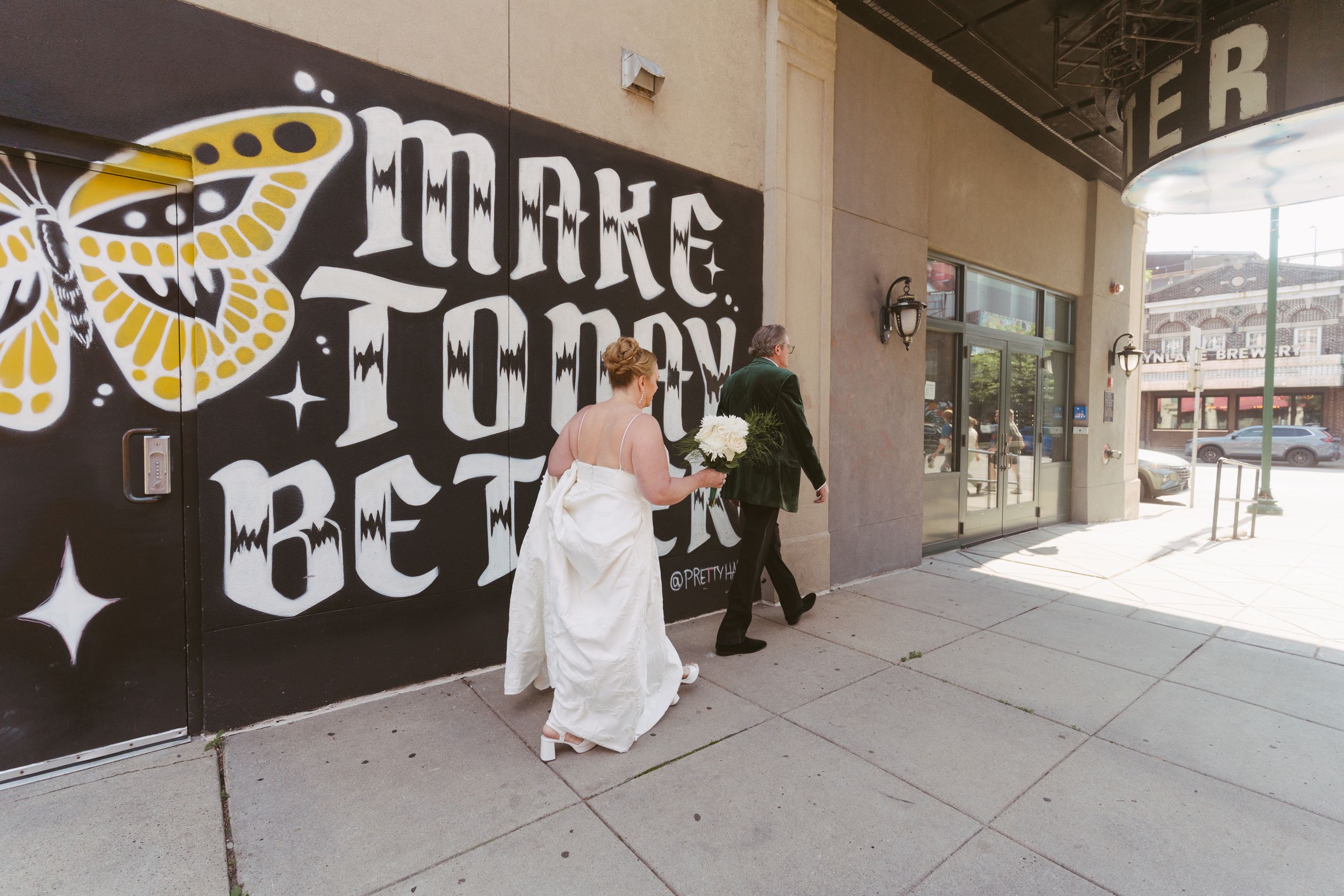 Bride and groom walk past a bold black mural reading Make Today Better on their wedding day.