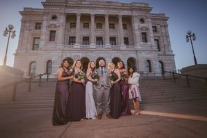 Groom poses with bridesmaids in purple gowns on steps of grand stone building at sunset wedding.