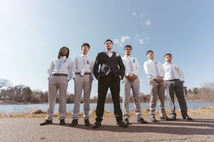 Groom in black suit stands confidently with groomsmen in white shirts and gray pants near a lake on a sunny day.