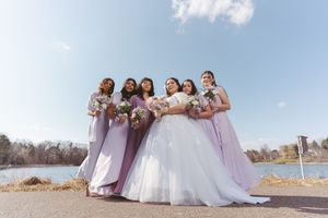 Bride in white ball gown poses with six bridesmaids in lavender dresses holding bouquets near a lake.