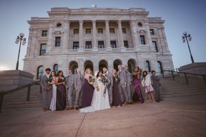 Wedding party poses on grand courthouse steps, bridesmaids in purple gowns and groomsmen in gray suits flanking bride and groom.