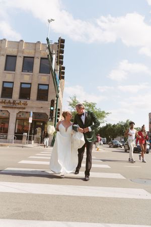 Bride and groom crossing a city street crosswalk in wedding attire on a sunny day.