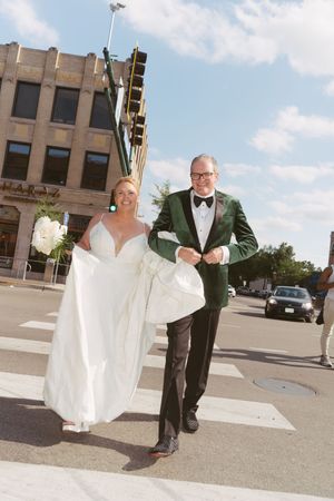 Smiling bride in white gown and groom in green velvet tuxedo cross a city street together on wedding day.