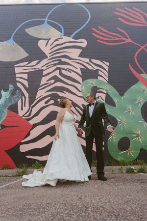 Wedding couple walks hand in hand before a bold street art mural with zebra and botanical designs.