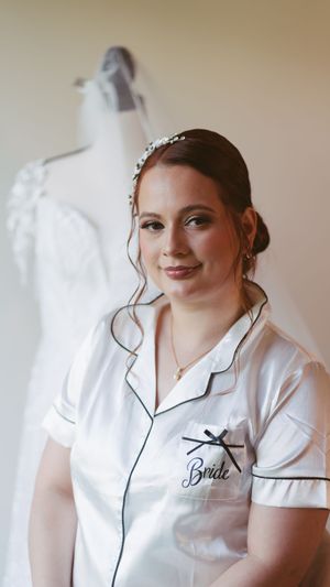 Bride wearing satin pajamas with floral headpiece, wedding dress on mannequin in background.