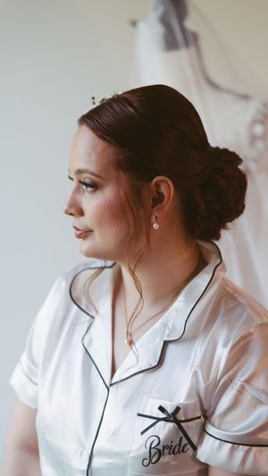 Bride in white satin pajamas with updo hairstyle and pearl earrings, wedding dress visible in background.