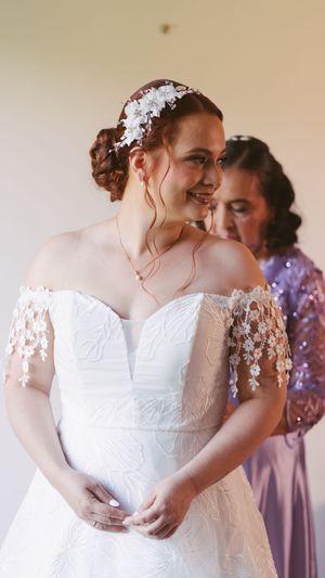 Smiling bride in off-shoulder lace wedding gown with floral headpiece being assisted by bridesmaid in lavender dress.