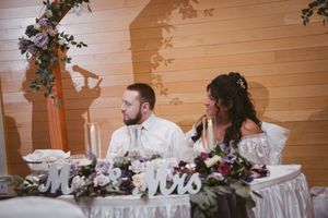 Bride and groom seated at sweetheart table decorated with purple florals and Mr. and Mrs. signs at wedding reception.
