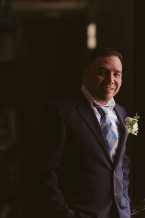 Smiling groom in navy suit with blue tie and white boutonniere poses in dramatic dark lighting.