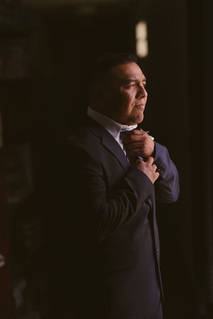 A groom in a navy suit adjusts his bow tie in dramatic lighting before his wedding ceremony.