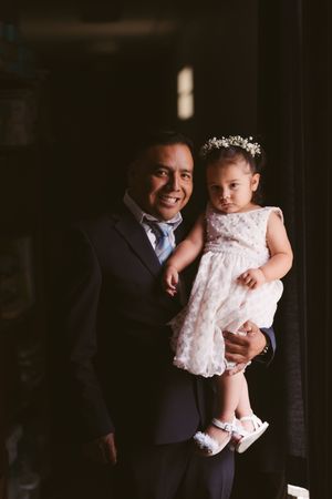 A smiling man in a navy suit holds a baby girl in a white lace dress with a floral headband.