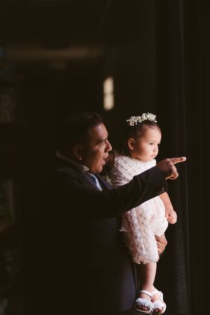 A groom in a navy suit holds a baby girl in a white dress and floral crown, pointing into the distance.