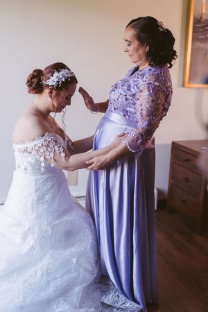 Bride in white lace gown being helped by woman in lavender dress during wedding preparations indoors.