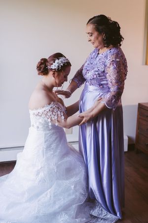 Bride in white lace gown with floral headpiece being helped by woman in lavender dress during wedding preparation.