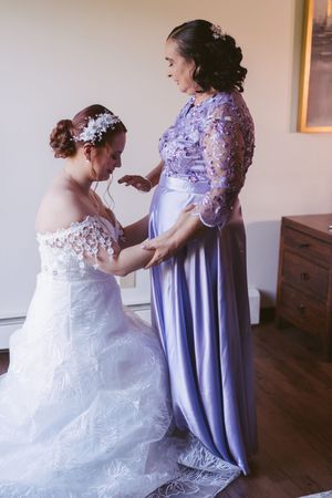 Bride in white lace gown holding hands with woman in lavender floral dress during wedding preparation.