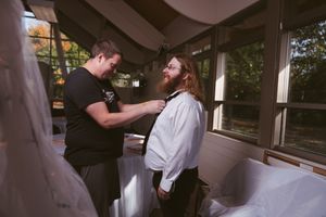 A man helps another adjust his tie while getting ready indoors, surrounded by soft natural light.
