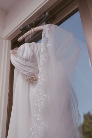 Elegant white wedding dress with lace veil hanging on a wooden hanger by a sunlit window.